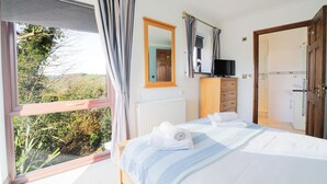 A bedroom with a large window, a bed with white linens, a wooden dresser, a mirror, and a television. - Nestled in an elevated position with sweeping views across the Luxulyan Valley. (Penpillick)