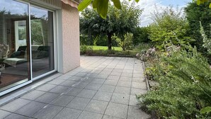 Eine Terrassenfläche mit Fliesenboden, eine Glastür, die zu einem Raum führt, und ein Garten mit verschiedenen Pflanzen und Bäumen.
