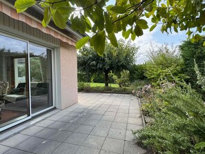 Eine Terrassenfläche mit Fliesenboden, eine Glastür, die zu einem Raum führt, und ein Garten mit verschiedenen Pflanzen und Bäumen.