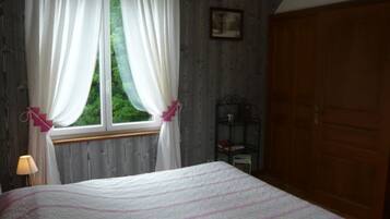 A bedroom with a bed, a window with curtains, a nightstand with a lamp, and a wooden wardrobe.