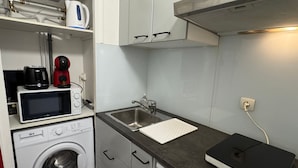 Een compacte keuken met een wasmachine, magnetron, koffiezetapparaat en een weegschaal op het aanrecht.