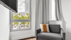 Ein modernes Wohnzimmer mit grauem Sofa, einem flachbildfernseher an der Wand und einem Fenster mit Blick auf Gebäude und Bäume.