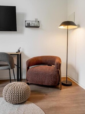 A modern living room with a brown upholstered armchair, a round knitted ottoman, a floor lamp, and a small table with decorative items. - Global Living Jena Rathausgasse (Jena)