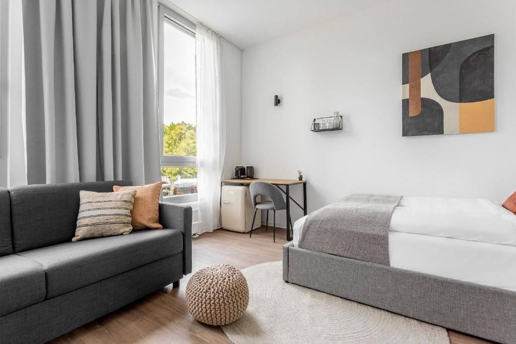 Ein modernes Hotelzimmer mit grauem Sofa, einem Bett, einem Schreibtisch mit Computer und einem Fenster mit Vorhängen.