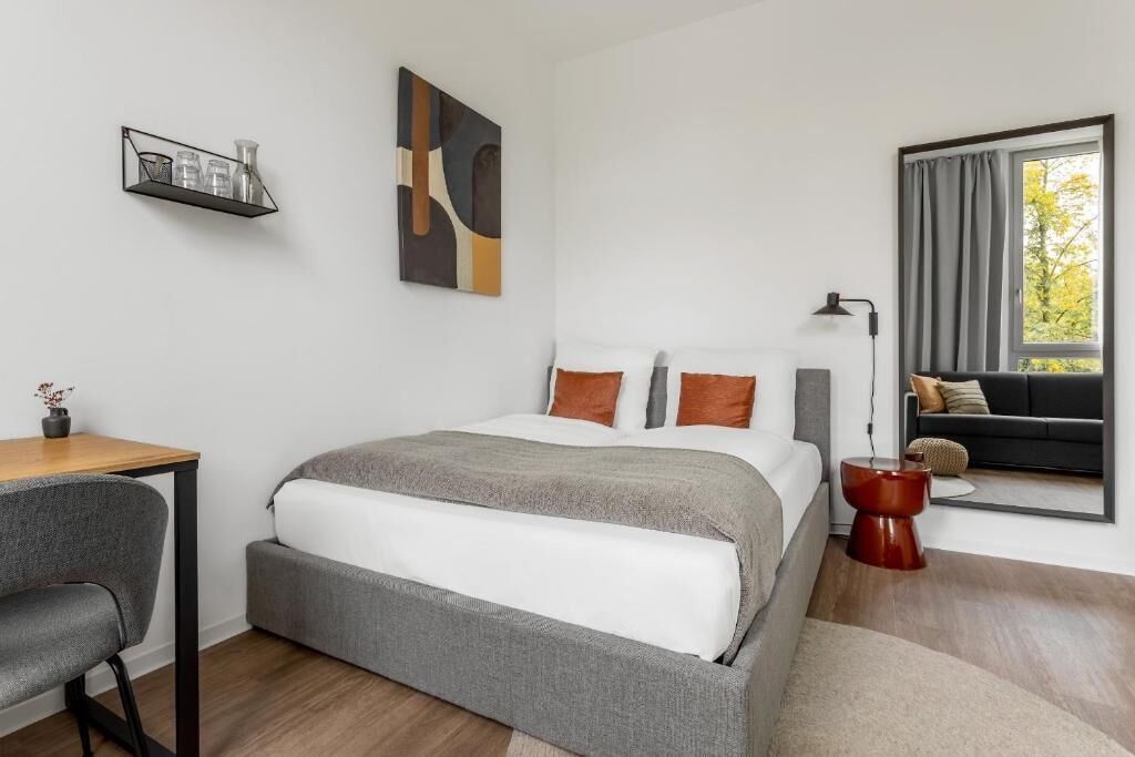 A modern bedroom with a bed, a desk, a chair, a side table, a lamp, and a window with curtains.