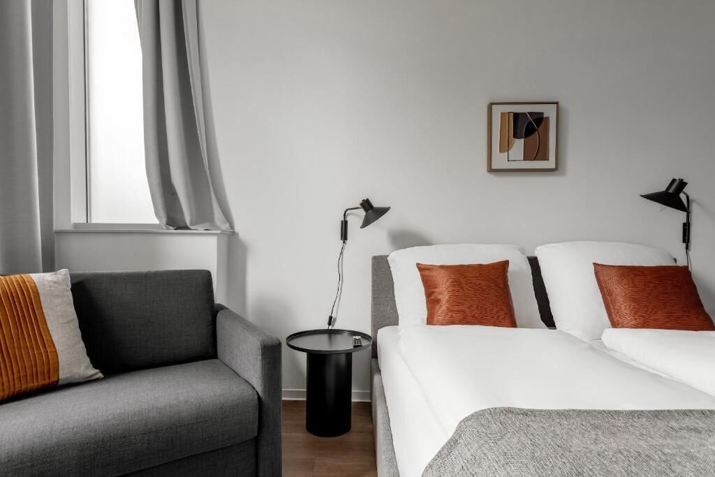 Una habitación de hotel moderna con un sofá gris, una cama con almohadillas blancas y anaranjadas, una mesita de noche negra pequeña, una lámpara de pie y una obra de arte abstracta enmarcada en la pared.