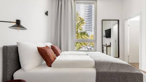 Ein modernes Schlafzimmer mit einem Bett, einem Fenster mit Blick auf ein hohes Gebäude, einem Schreibtisch und einem Stuhl.