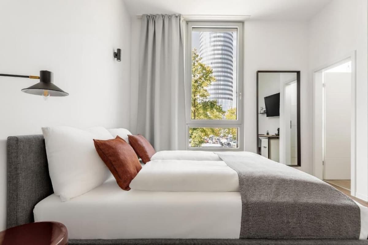 A modern bedroom with a bed, a window with a view of a tall building, a desk, and a chair.
