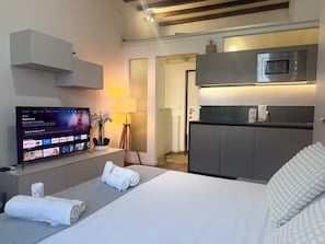 A modern hotel room with a bed, a TV displaying a streaming service, a small kitchenette, and a bedside table with a lamp and flowers. - ACasaDiImma Vicino Metro Wi-Fi Luminoso (Milan)