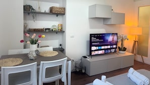 A modern living room with a TV displaying a streaming service interface, a dining area with a table and chairs, and a cozy seating area with a lamp and a potted plant. - ACasaDiImma Vicino Metro Wi-Fi Luminoso (Milan)