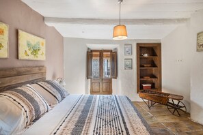 A bedroom with a bed, a wooden headboard, a hanging lamp, a wooden shelf with dishes, and a vintage wheelbarrow.