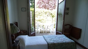 Una habitación con dos camas, un escritorio, una silla y un ventanal corredero que da a un espacio al aire libre con sillas.