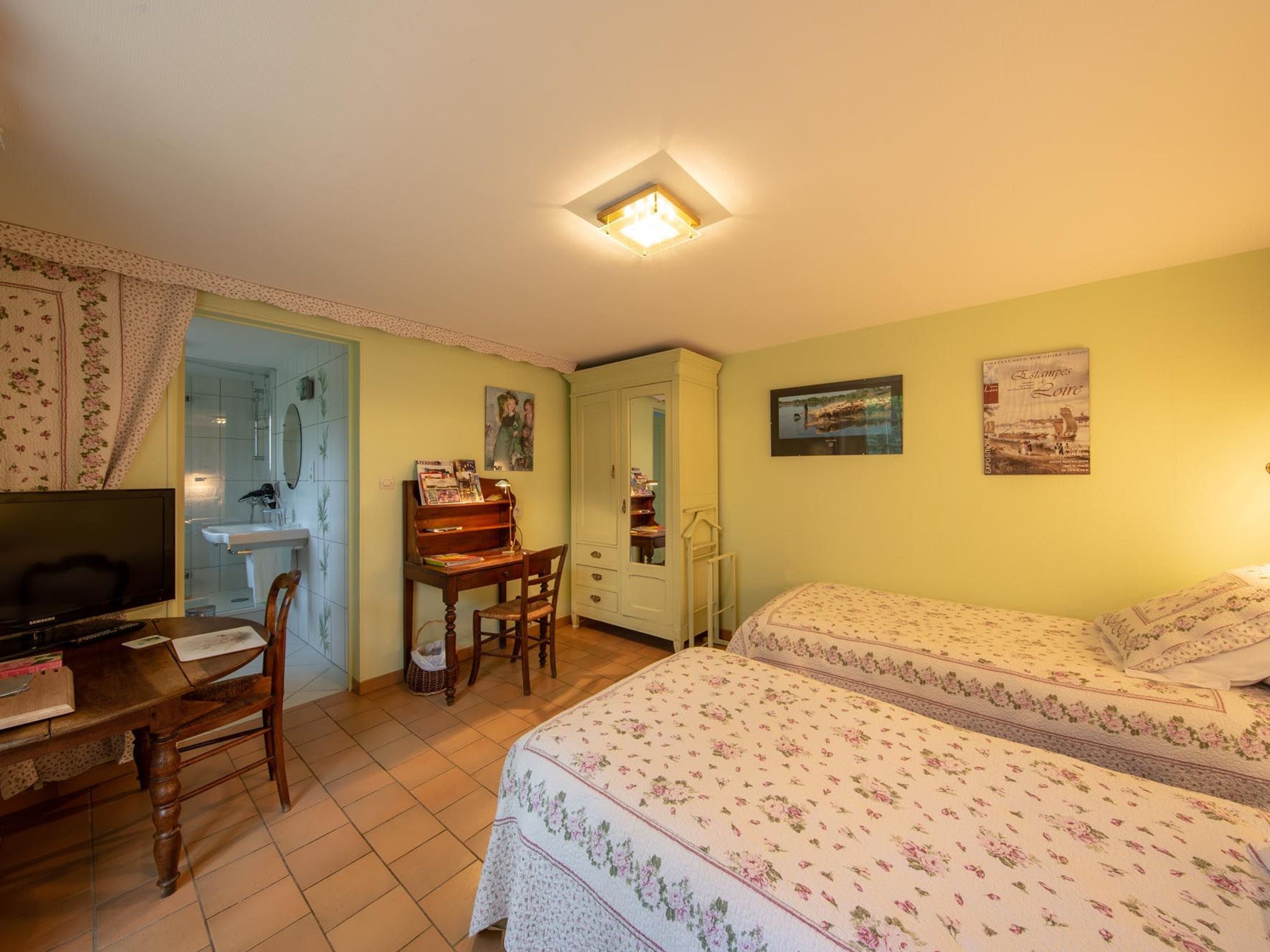 Une chambre à coucher avec un lit, un bureau avec une chaise, une télévision et une salle de bain visible à travers une porte ouverte.