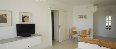 A modern living room with a flat-screen TV on a white cabinet, a white sofa, a dining area with a white table and orange chairs, and a white ceiling with pendant lights.