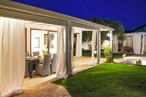 An outdoor covered patio area with wicker furniture and a dining table, surrounded by white curtains and a well-maintained lawn. - Villa Erika SicilyMyHome (Ispica)
