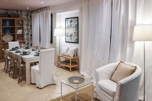 A dining area with a table set for four, a white armchair, a side table with a lamp, and a cabinet with dishes. - Villa Erika SicilyMyHome (Ispica)