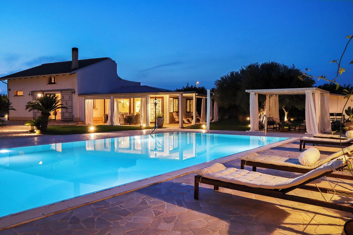 Une piscine avec des chaises longues, un patio couvert et une maison dont l’intérieur est éclairé.
