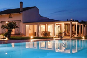 A poolside area with a covered seating area, a pool, and a house with a white facade and a dark roof. - Villa Erika SicilyMyHome (Ispica)
