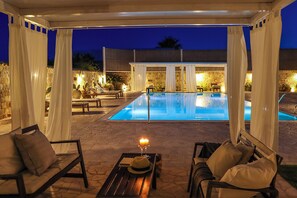 An outdoor pool area with a covered seating area, stone flooring, and ambient lighting. - Villa Erika SicilyMyHome (Ispica)