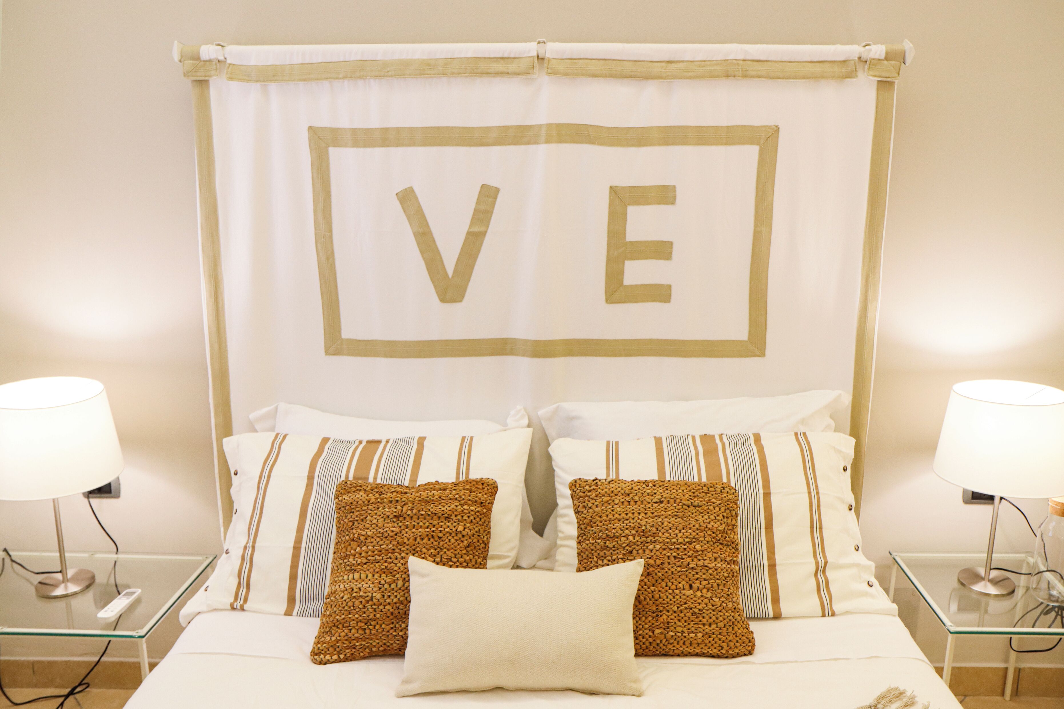 A neatly made bed with a large 'VE' sign on the headboard, two bedside lamps, and a glass side table.