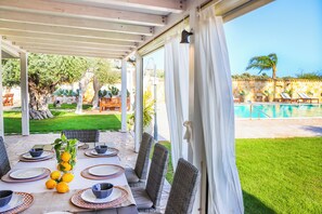 An outdoor patio with a table set for dining, surrounded by wicker chairs, a pool in the background, and a view of a garden and pool area. - Villa Erika SicilyMyHome (Ispica)