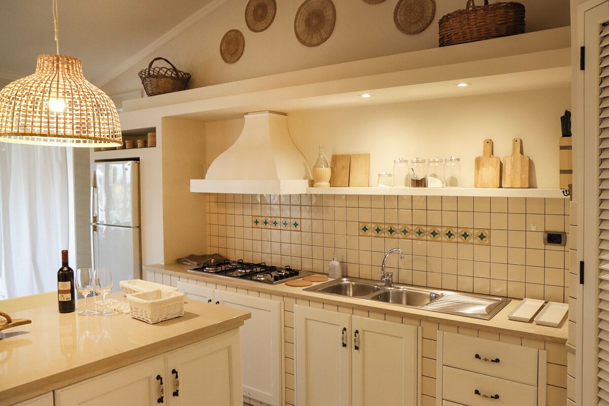 Une cuisine équipée d’une cuisinière à gaz, d’un évier et de meubles blancs. On y trouve également un luminaire suspendu et un panier fixé au mur.