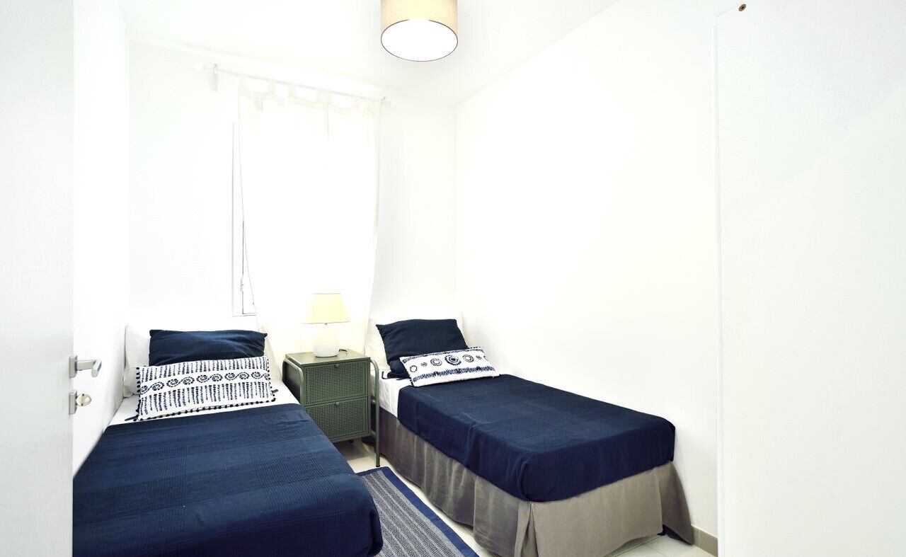 Une petite chambre propre, avec deux lits simples, une table de chevet, une lampe et une fenêtre avec des rideaux.