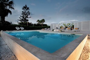 A rectangular swimming pool with a clear blue water surface, surrounded by a low stone wall. There are lounge chairs and umbrellas in the background, suggesting a resort setting. - Villa Elysian con Piscina Privata (Pachino)