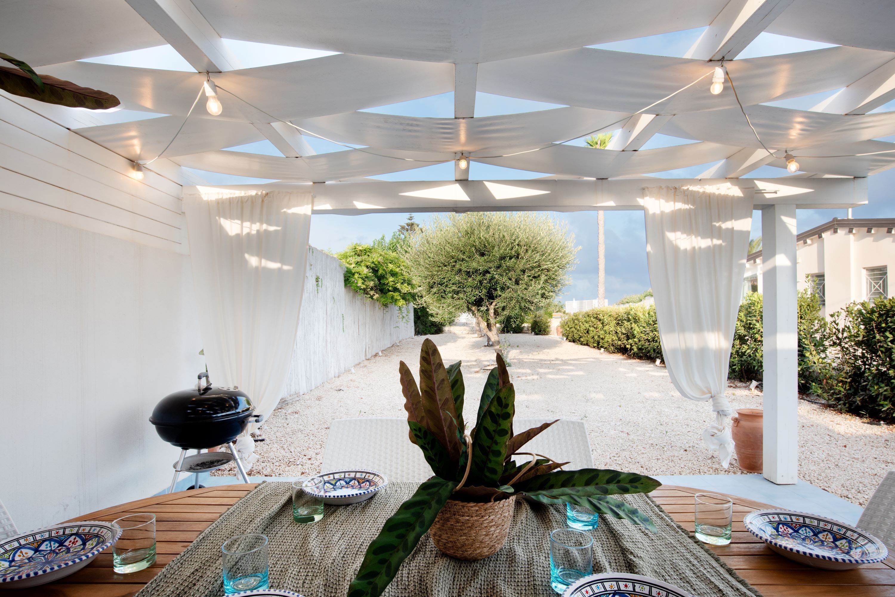 A covered outdoor dining area with a table set for two, a grill, and a potted plant.