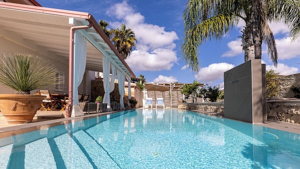 A clear swimming pool with a covered area and lounge chairs, surrounded by palm trees and a stone wall. - Villa Zamia SicilyMyHome      (Ispica)