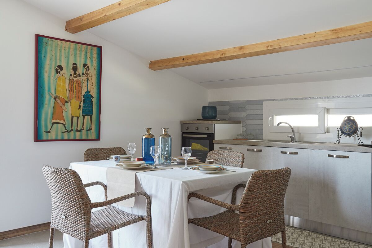 A dining area with a table set for four, surrounded by wicker chairs. A kitchen with white cabinets and a tiled backsplash is visible in the background.
