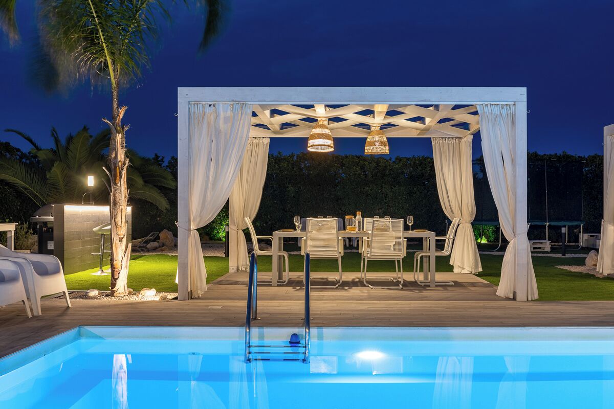 Un espace piscine doté d’un coin salon couvert, de mobilier blanc et d’une pergola agrémentée de tissus drapés.
