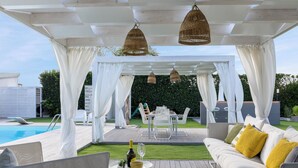 Une terrasse extérieure avec une piscine, des meubles blancs et une pergola agrémentée de guirlandes lumineuses.