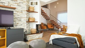 Un salon moderne avec un mur en pierre, un téléviseur à écran plat, un canapé, des poufs et un escalier menant à un espace repas avec des chaises.