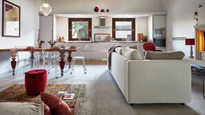 A modern living room with a sofa, a dining area with chairs, a kitchen in the background, and a large window. - Villa Scarlet con Piscina Privata (Modica)