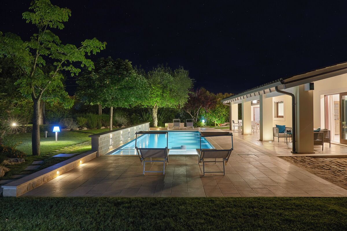A swimming pool with two lounge chairs, surrounded by a patio area with a covered seating area and a well-lit garden.