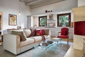 A modern living room with a sofa, coffee table, and red armchair. A kitchen with a window and a red chair is visible in the background. - Villa Scarlet con Piscina Privata (Modica)