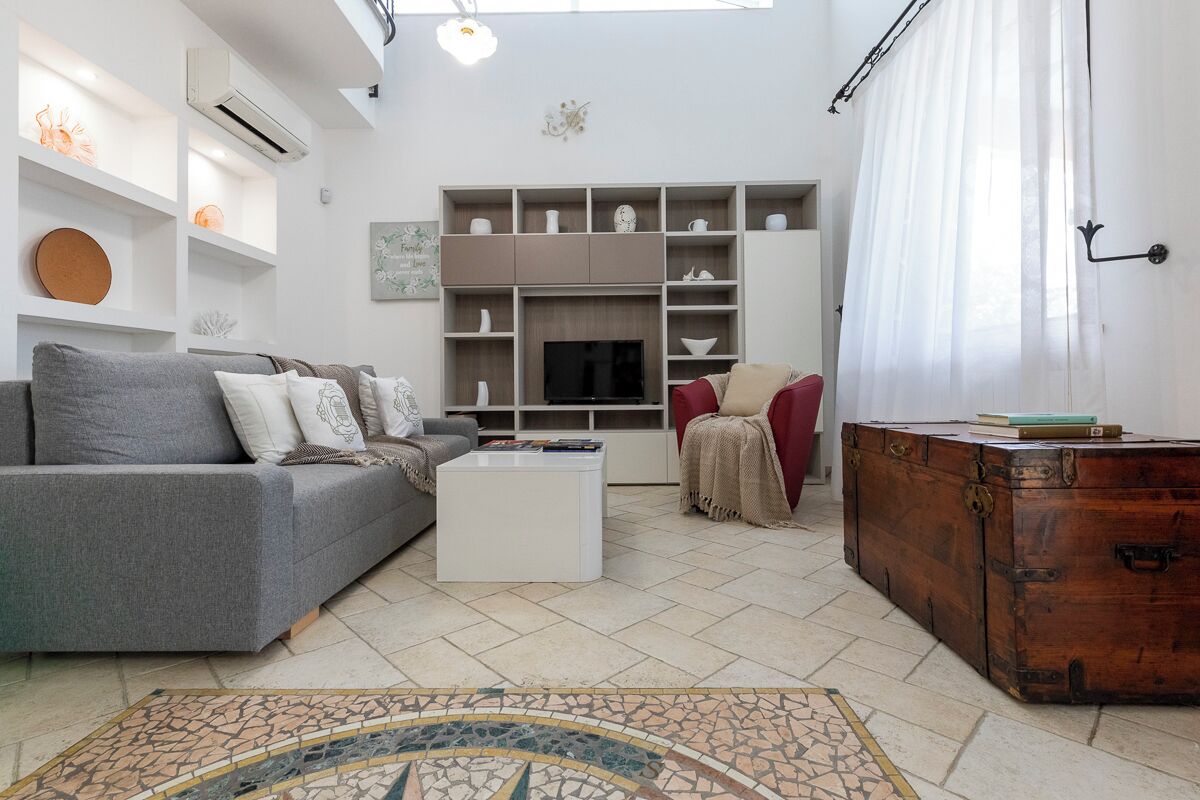 Ein Wohnzimmer mit einer Couch, einem Couchtisch und einer Holzkiste. Es sind außerdem Regale mit Dekoration und ein Fernseher zu sehen.
