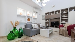 A living room with a grey sofa, white coffee table, and green vases. Shelves with decorative items and a flat-screen TV are also visible. - Villa del Colle con Piscina Privata (Ispica)