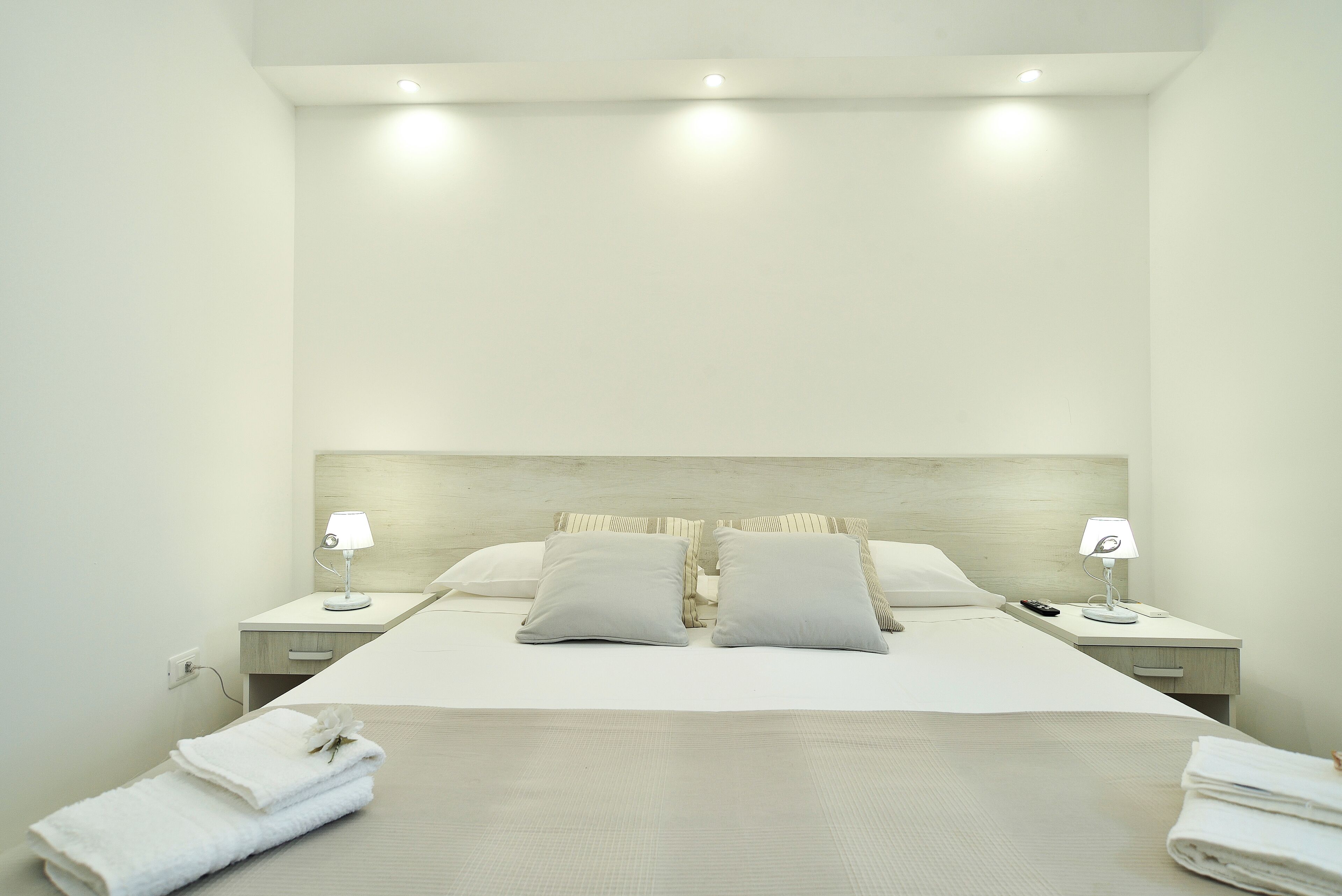 A neatly made bed with two pillows, a bedside table with a lamp, and a folded towel on the floor.