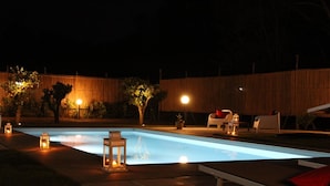 Piscina con cubierta de baldosas, rodeada por una cerca de madera y árboles. Hay sillas de descanso y una pequeña mesa con velas.