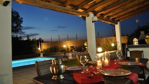 Un área de comedor al aire libre con piscina, pérgola de madera e iluminación nocturna.