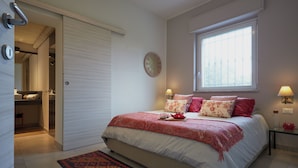 Un dormitorio con cama, ventana, mesita de noche y acceso a un baño.