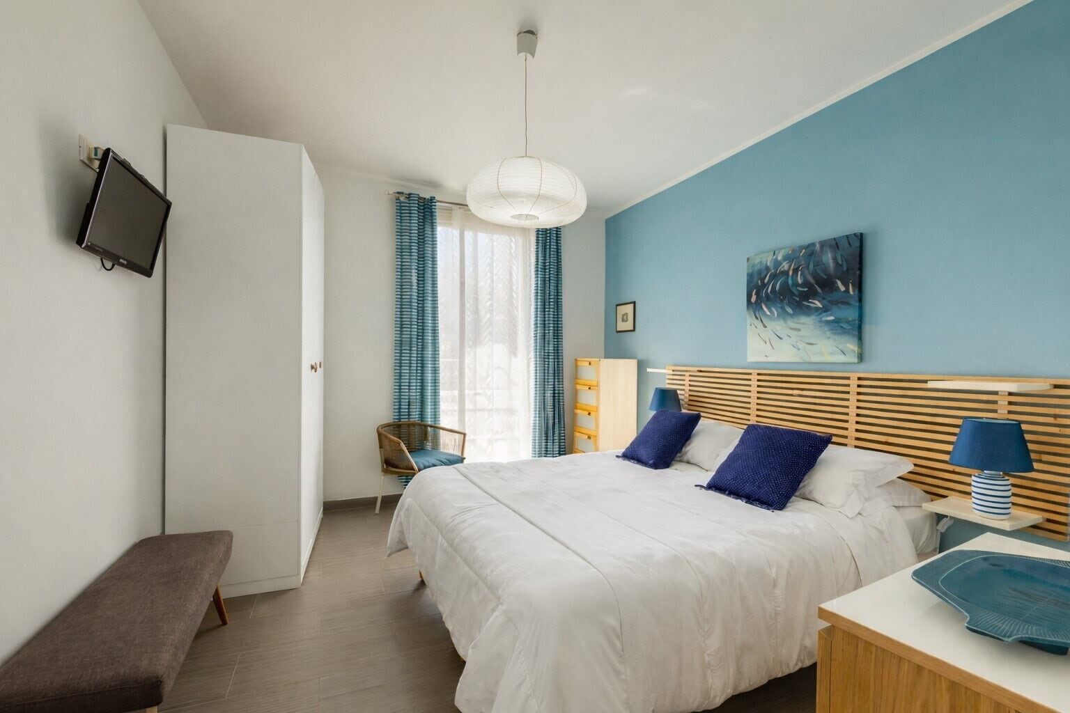 A bedroom with a bed, bedside table, lamp, chair, and a television on the wall.