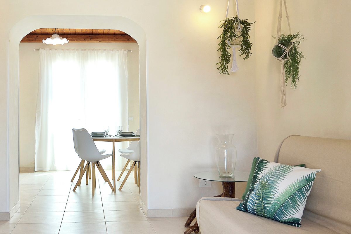 Zona pranzo con tavolo e sedie, un tavolo rotondo in vetro, una poltrona con cuscino a motivo di foglie verdi e una pianta pensile.