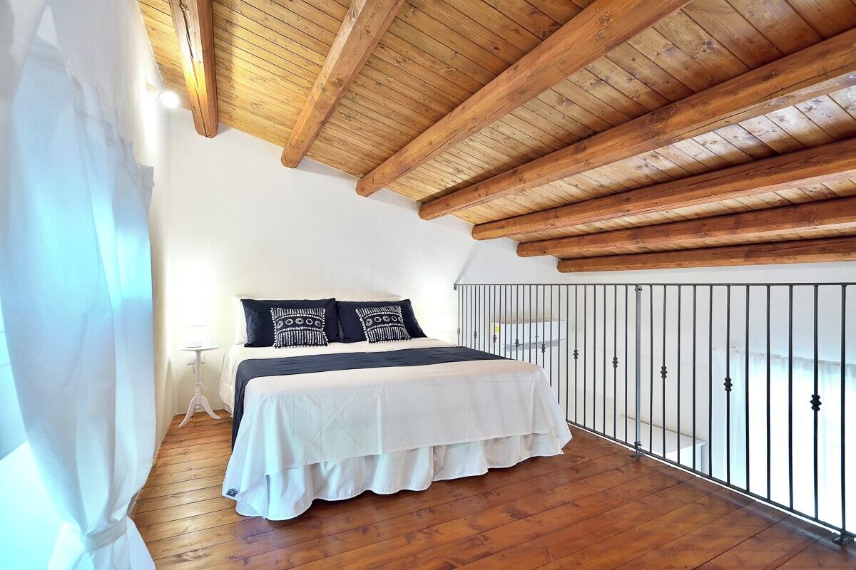 Een slaapkamer met een houten plafond, een bed met witte beddengoed en een metalen leuning bij het raam.