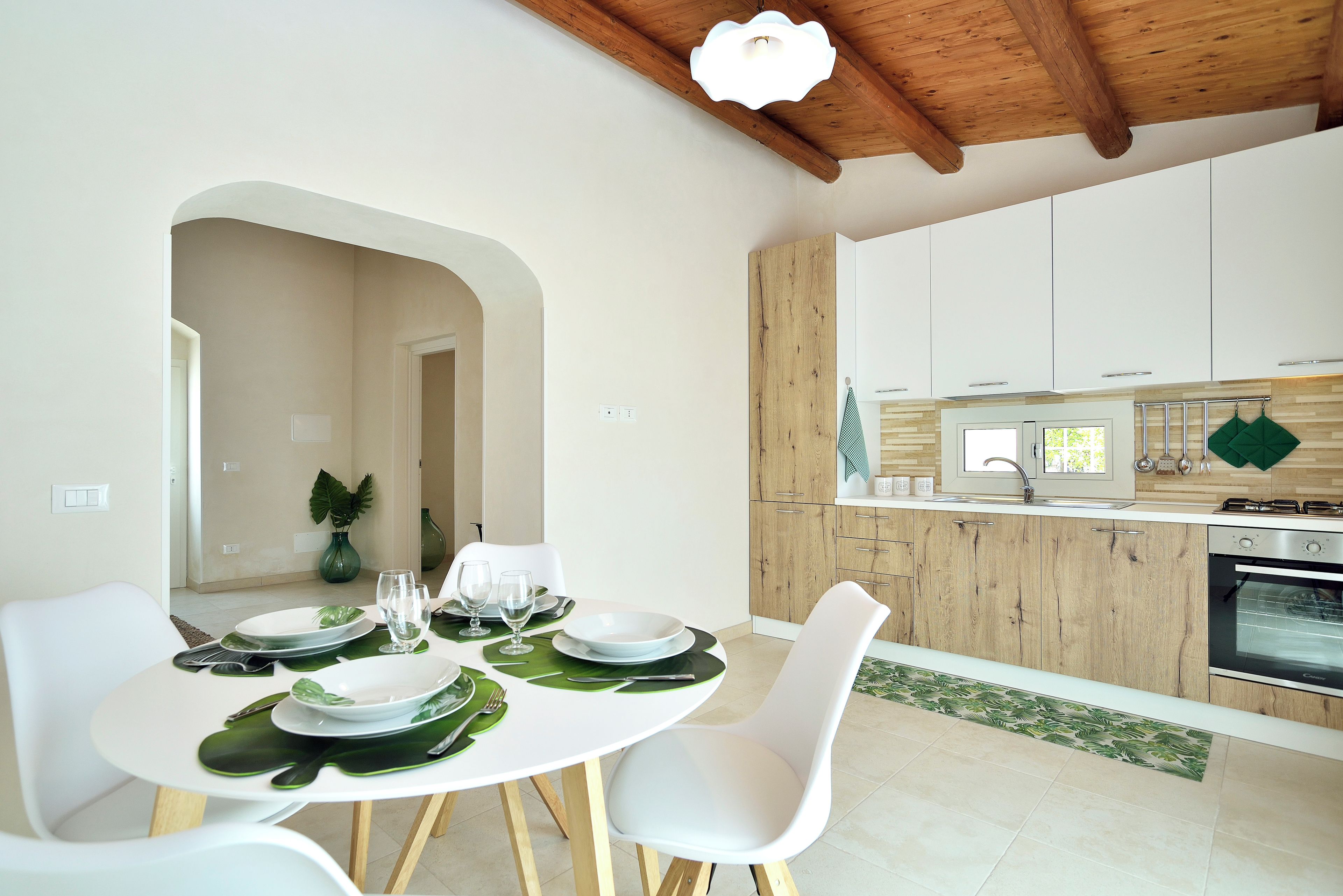 A modern dining area with a white table set for four, a kitchen with wooden cabinets, and a view of the dining area through an archway.