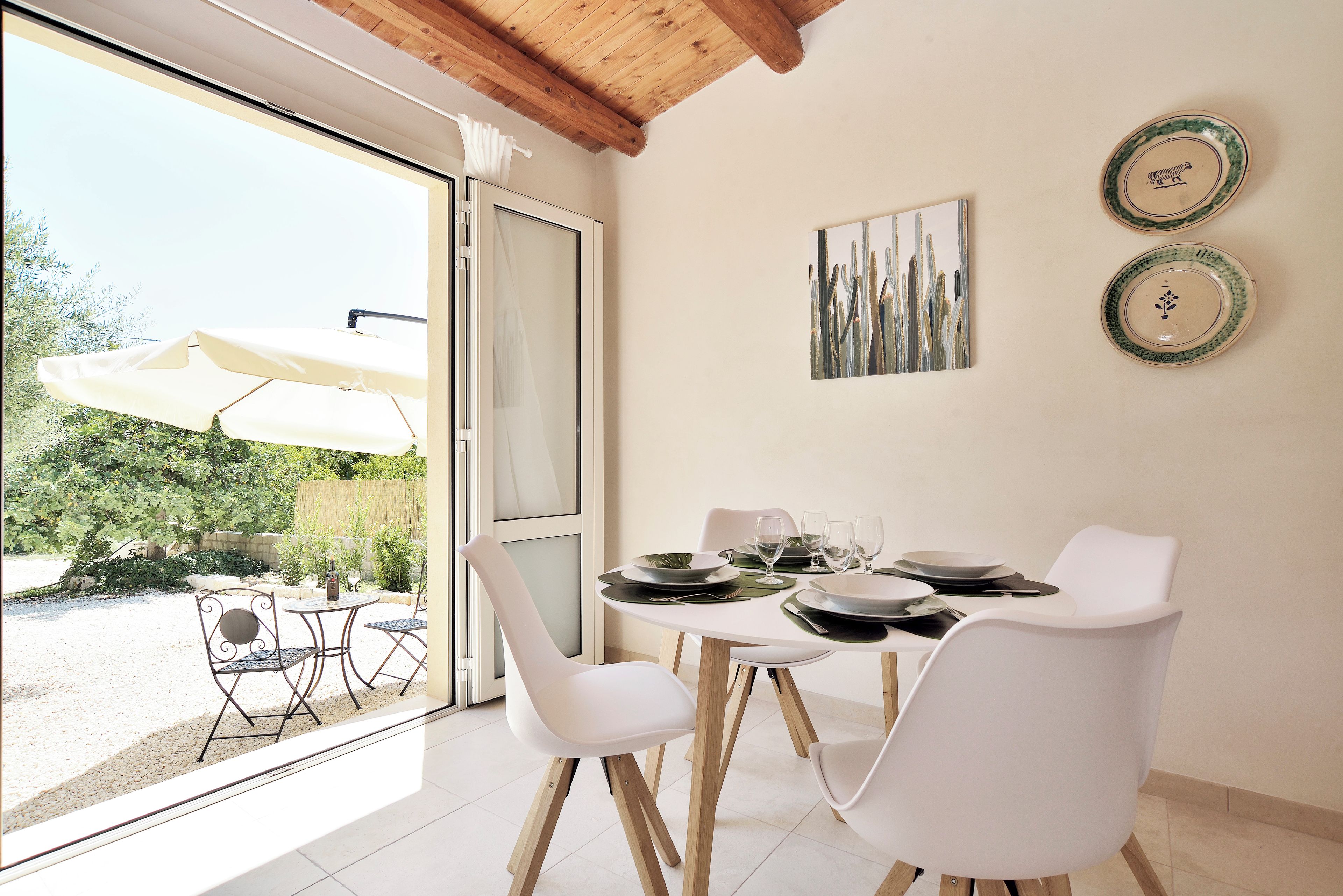 Zona pranzo con tavolo apparecchiato per quattro, sedie bianche e vista sul giardino tramite una porta in vetro.