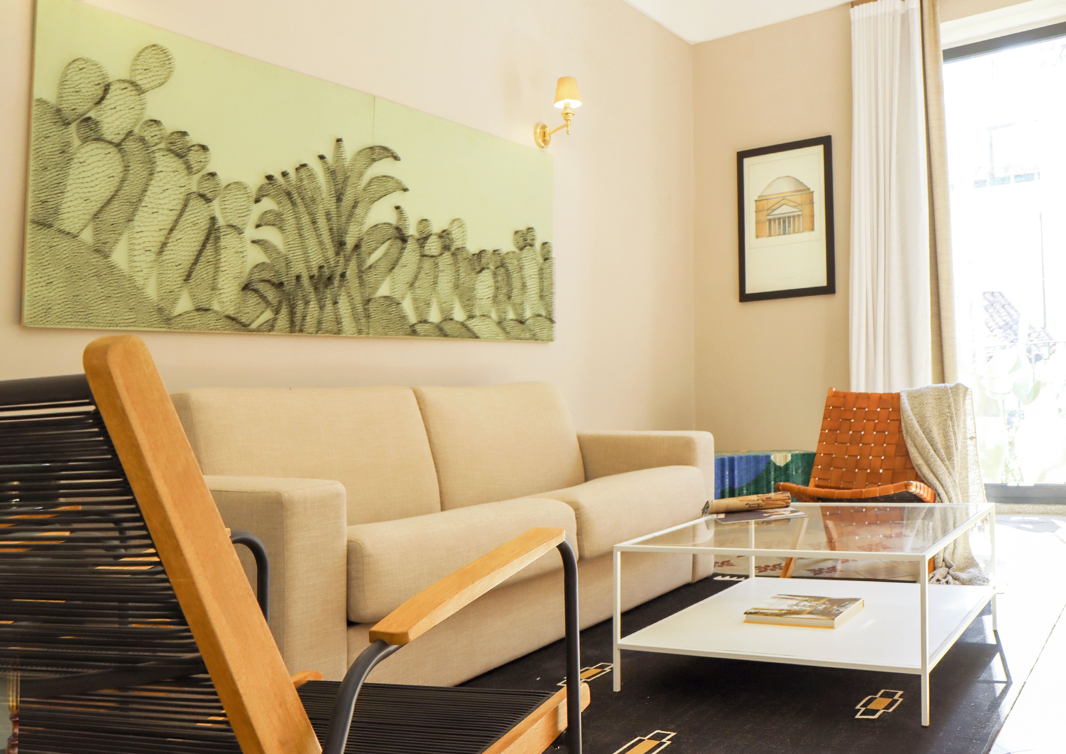 A living room with a beige sofa, a wooden chair, a glass coffee table, and a cactus artwork on the wall.