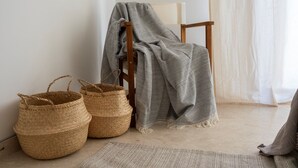 Una habitación con una silla de madera cubierta por una manta gris, dos cestas tejidas y una alfombra color beige.